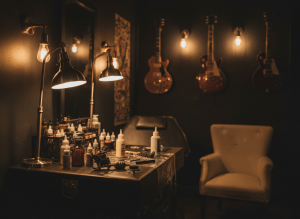 Moody, atmospheric photograph of a dark tattoo studio interior with warm brass and gold accent lighting, near-black walls, vintage rock and roll aesthetic, guitars faintly in the background, a tattoo machine and ink bottles on a dark leather station, cinematic low-key lighting, deep shadows, cream and gold tones, no text, no visible faces