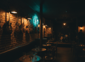 Moody, atmospheric photograph of a dimly lit music venue interior at night. Near-black darkness with warm brass and amber lighting glows, vintage guitars on a wall, exposed brick, a hint of neon. Cinematic, rock-and-roll dive-bar mood, deep shadows, warm gold highlights. No text, no people faces.