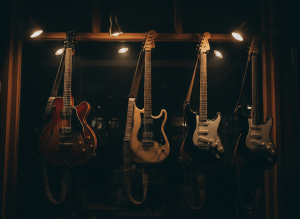 Dark atmospheric photograph of vintage electric guitars hanging in a dimly lit independent guitar shop window, warm amber and brass lighting, deep shadows, moody near-black background, no bright whites